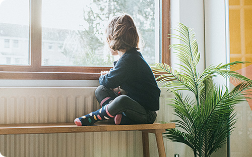 Kleiner Bub schaut beim Fenster raus. Neben ihm eine Pflanze.